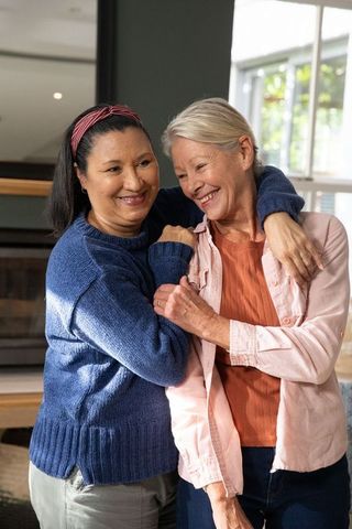 Mature women hugging and laughing in cozy living room with fireplace mantel and sunlit window