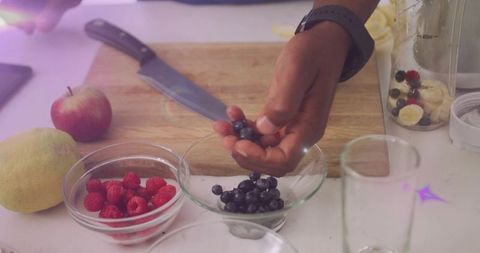 Preparing fresh berry smoothie ingredients with hands placing blueberries into glass bowl