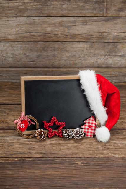 Santa hat and various Christmas decorations arranged on a slate with a rustic wooden background. Ideal for holiday greeting cards, festive invitations, seasonal advertisements, and Christmas-themed social media posts.