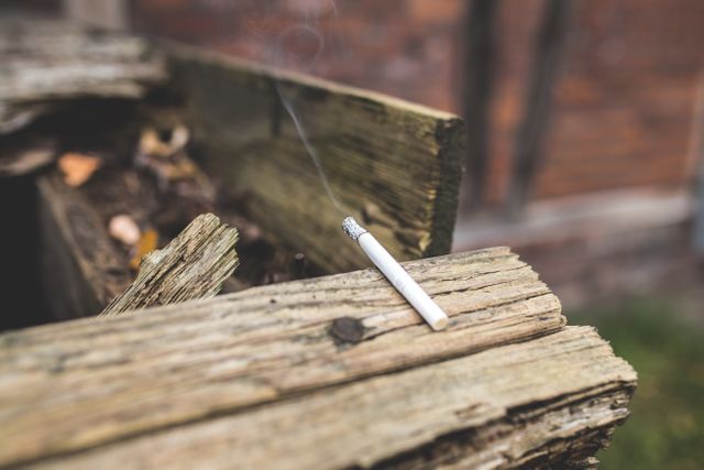 A close-up shot of a cigarette resting on a weathered wooden surface in an urban setting with smoke rising. Ideal for use in campaigns and articles addressing smoking, health risks, urban life, or environmental concerns. Can also be used in public health awareness materials, advertisements for smoking cessation, or content related to lifestyle choices.
