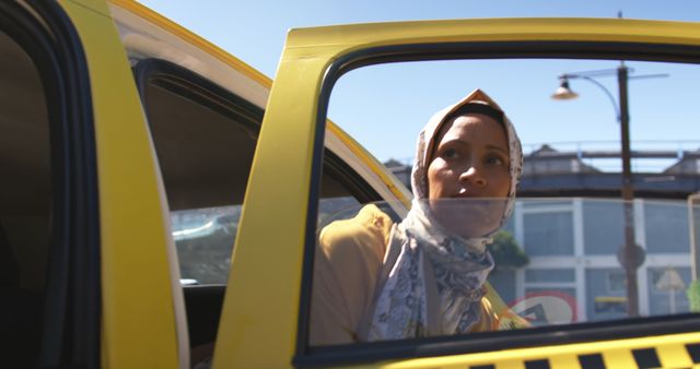 This photo depicts a Muslim woman wearing a headscarf, in the process of exiting a yellow taxi on a sunny day. The urban background suggests modern city life and routine commuting. This image can be used to promote topics related to travel, multiculturalism, daily commute, city life, and religious diversity.
