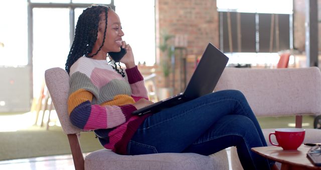 Smiling woman sitting comfortably while talking on the phone and using a laptop. Ideal for use in articles or posts about remote work, home office setup, productivity, and modern lifestyle.