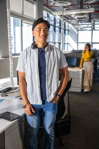 Young Asian man standing in sunlit modern office wearing headphones, coworker speaking on phone