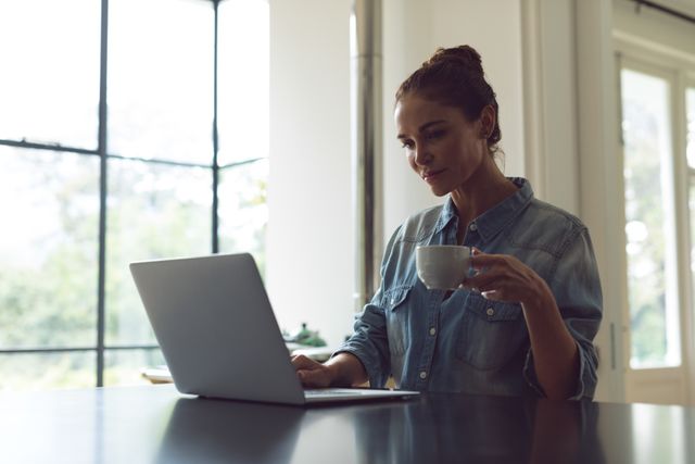Front view of woman drinking coffee while using laptop on worktop in kitchen at comfortable home
