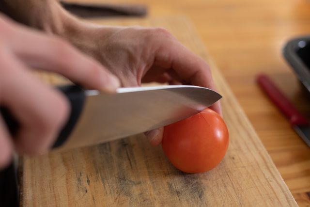 Caucasian female chef slicing a fresh tomato on a wooden chopping board in a professional kitchen. Ideal for use in articles or advertisements related to culinary arts, professional cooking, restaurant kitchens, food preparation, and healthy eating.
