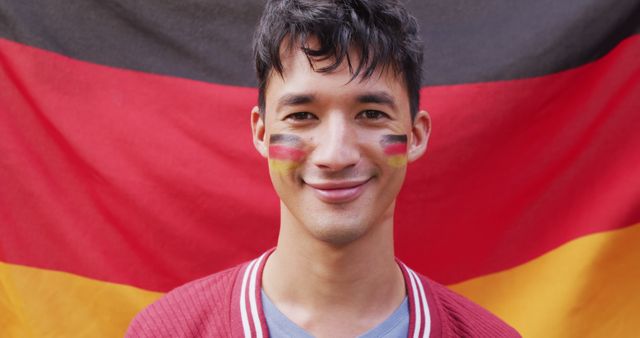Young male fan smiling, face painted with German flag colors, standing in front of large German flag. Image conveys pride, patriotism, and support. Ideal for use in sports promotions, fan pride advertisements, cultural events or festivals.
