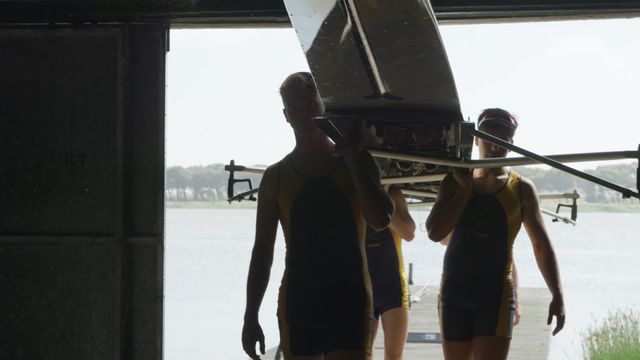 Team of male rowers effortlessly carrying a rowing boat into a boathouse. The image showcases teamwork and determination in rowing sports, perfect for illustrating themes of collaboration, outdoor athletics, or athletic training. Useful for sports-related articles, team spirit promotions, and outdoor sporting event advertisements.