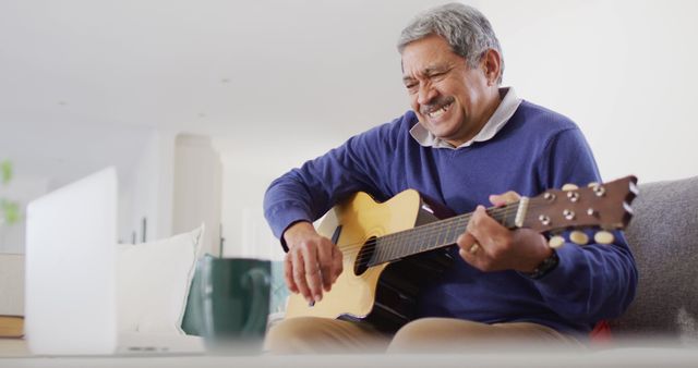 Image of smiling senior biracial man using laptop and playing acoustic guitar at home. Retirement, hobbies, global communication, domestic life, inclusivity and senior lifestyle concept.