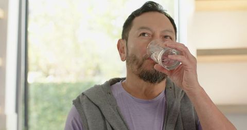 Mature Asian Man Drinking Water Near Sunlit Garden Window