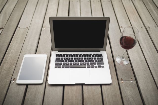 High angle view of a laptop, digital tablet, and wineglass placed on a wooden porch. Ideal for illustrating remote work, modern lifestyle, or relaxation themes. Suitable for articles, blogs, or advertisements focusing on technology, work from home setups, or leisure activities.