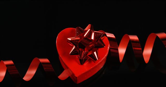 A red heart-shaped box adorned with a shiny bow is complemented by a curling ribbon against a dark background, with copy space. Perfect for conveying themes of love, Valentine's Day, or romantic celebrations.
