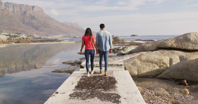Diverse couple walking on pier and holding hands with copy space by sea. Outdoors, summer, vacations, relaxing, free time, love and togetherness, unaltered.