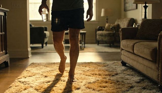 Barefoot man walking across sunlit living room shag rug holding glass of water