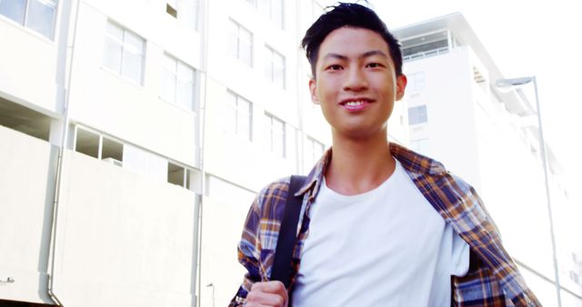 A young Asian male student smiles as he walks outdoors on a campus, with copy space. His casual attire and backpack suggest a relaxed university environment.