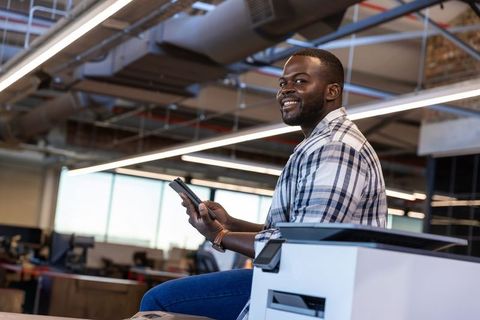 Smiling african american professional sitting on office printer using smartphone for work