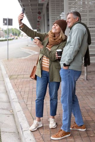 Mature couple taking selfie with coffee at urban transit shelter, autumn layers