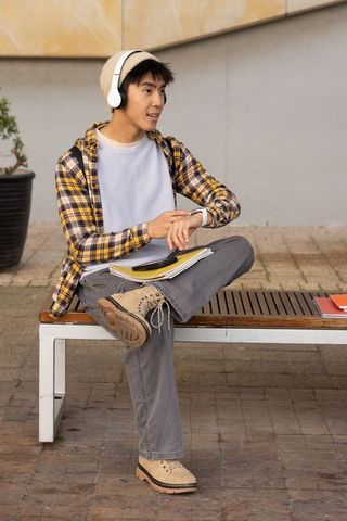 Asian teen student listening to headphones and checking smartwatch on campus bench