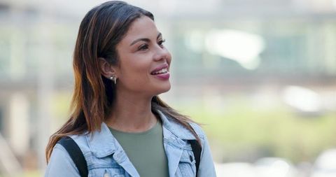 Smiling young woman looking upward in urban plaza wearing denim jacket and backpack