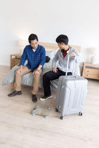 Asian men sitting on bed edge with suitcase in minimalist bedroom preparing to travel