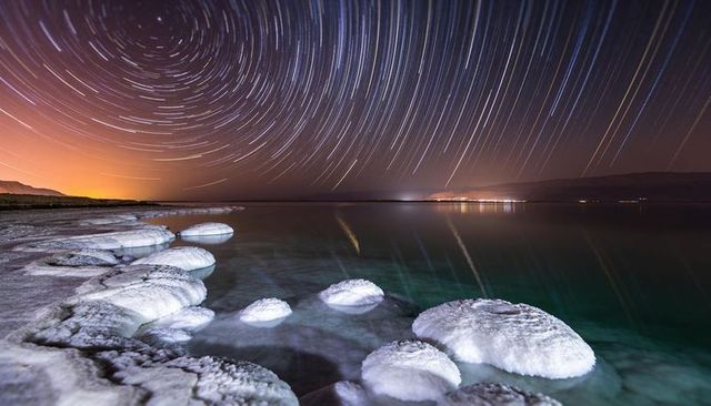 Glowing salt mounds along dead sea shore under swirling star trails and teal reflections