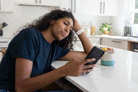 African american woman sitting at kitchen island leaning on hand while using smartphone