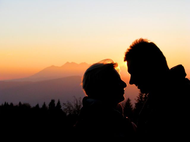 Romantic couple facing each other silhouetted against an orange sunset backdrop with distant mountains. Perfect for illustrating love, romance, serenity, evening tranquility. Suitable for use in travel brochures, romantic greeting cards, relationship blogs, and nature websites.