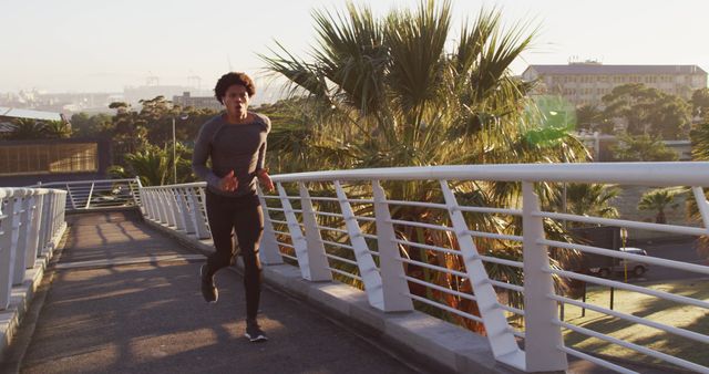 A young man is jogging on an outdoor bridge path during sunset. He is dressed in casual running attire and appears focused on his exercise routine. Palm trees and a distant urban area provide a scenic backdrop. Ideal for use in fitness, lifestyle, or wellness promotions, as well as advertisements encouraging outdoor activities and healthy living.