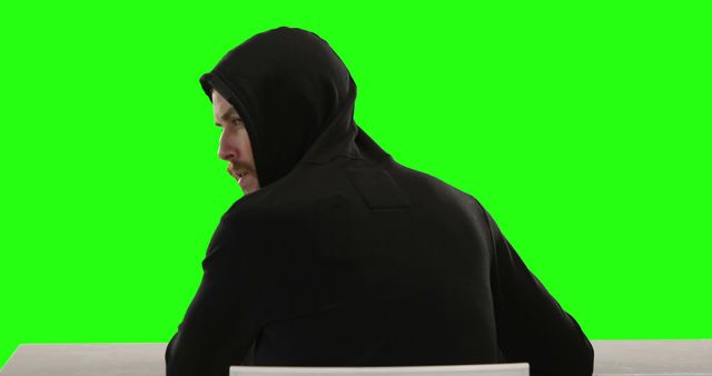 Hooded man facing away from camera with green screen background seated at table. Perfect for visual effects, advertising layouts, scene incorporation in films, tutorials on using a green screen.