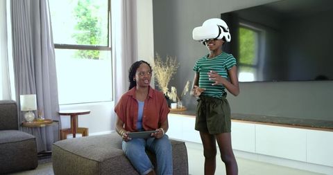 African American mother guiding teenage daughter using virtual reality headset at home