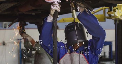 Automotive welder repairing car undercarriage wearing helmet, apron and coveralls
