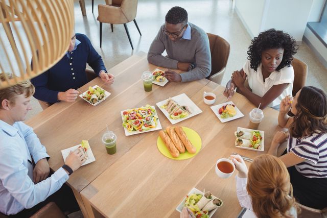 This image shows a diverse group of business coworkers having a breakfast meeting around a table in an office cafeteria. They are enjoying a variety of food items including sandwiches, salads, pastries, and beverages. Perfect for illustrating teamwork, corporate culture, casual workplace meetings, and diverse work environments.