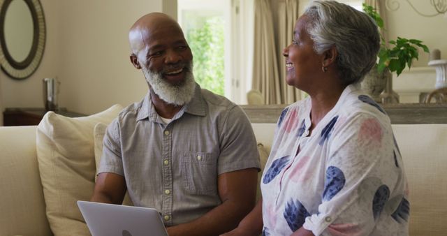 African american senior couple looking at each other and smiling while using laptop together at home. retirement senior couple lifestyle living concept