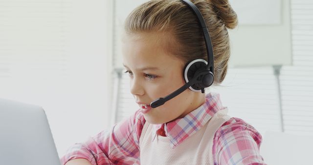 A young Caucasian girl is focused on her computer screen while wearing a headset, with copy space. Her engagement suggests she might be participating in an online class or virtual communication.