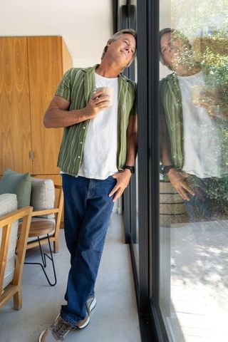 Mature man leaning by glass sliding door holding coffee mug, relaxed morning lifestyle