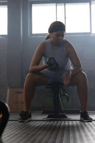 African american woman resting on bench lifting dumbbell in industrial gym backlight