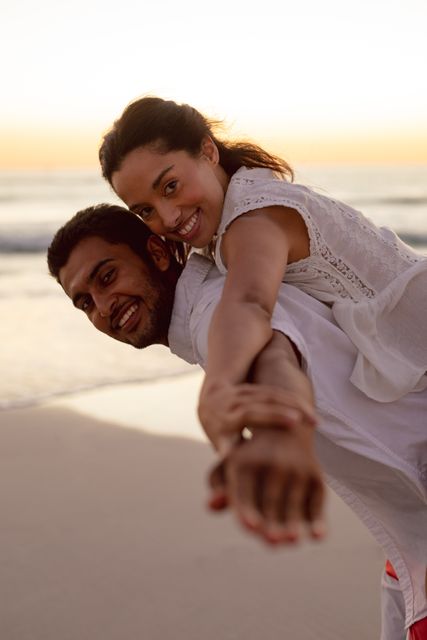 This image captures a joyful couple enjoying a playful piggyback ride on the beach during sunset. Perfect for use in travel brochures, romantic getaway promotions, lifestyle blogs, and advertisements focusing on love, happiness, and outdoor activities.