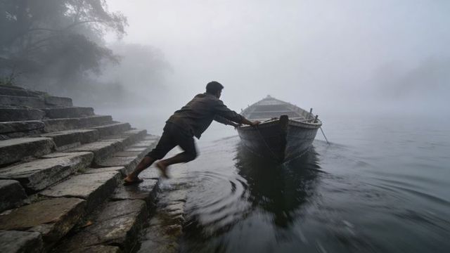 Boatman pushing wooden rowboat from stone ghats at foggy dawn mist