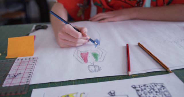 Designer drawing fashion sketches using colored pencils on desk. Ideal for content about creativity, fashion design, artistic process, and creative workspaces.