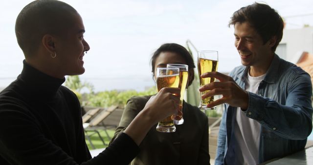 Three friends enjoying each other's company while clinking glasses of beer in a cheerful outdoor setting. Ideal for use in advertising campaigns, social media content, articles, and blog posts focusing on celebration, leisure activities, and social gatherings.