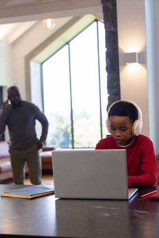 African american father supervising child studying at home with laptop and headphones