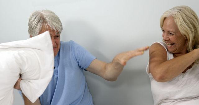 Senior couple seen in playful pillow fight, expressing joy and laughter. Can be used in campaigns promoting active senior lifestyle, happiness in elderly relationships, family bonding activities, and positive aging.