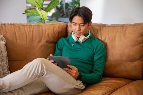 Asian man relaxing on tan leather sofa using tablet with headphones and smartwatch