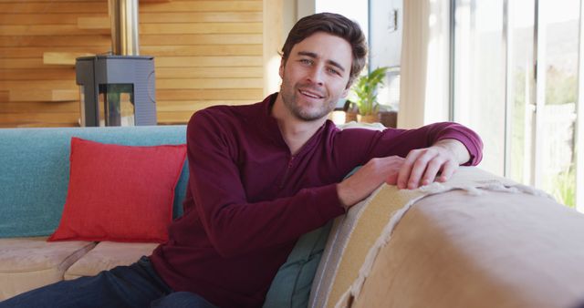 A man is seen smiling while sitting comfortably on a cozy sofa in a modern living room. This scene exudes warmth and relaxation, perfect for themes related to home decor, comfort, casual lifestyle, and indoor leisure activities. Ideal for use in advertisements, blog posts about home design, and articles related to relaxation and mental well-being.