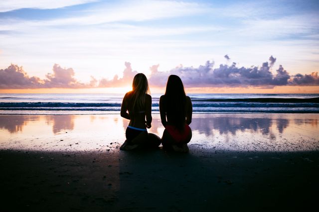 Two women are seen sitting on a beach during sunset, creating a peaceful and serene atmosphere as the sky and sea reflect beautiful colors. This can be used for themes relating to relaxation, friendship, travel, adventure, and the tranquility of nature.