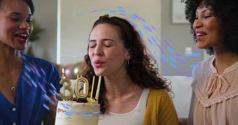 Blowing 30th birthday candles on frosted two-tier cake while friends celebrating on cozy sofa