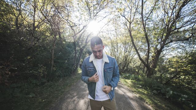 Young man wearing denim jacket walking on sunlit forest path surrounded by lush green trees. Perfect for illustrating outdoor activities, nature exploration, tranquility, and casual fashion concepts. Can be used for travel blogs, fashion advertisements, and lifestyle articles. Represents peaceful, mindful moments alone in nature.