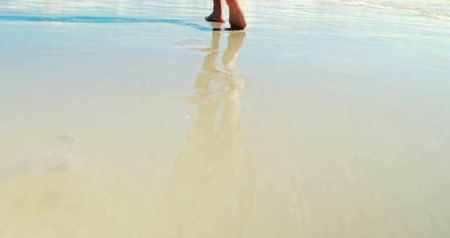 A person is seen walking barefoot on wet beach sand, casting a reflection on the water, during a sunny day. Suitable for use in advertisements for vacation destinations, beach outfits, or travel blogs. This image evokes feelings of relaxation and tranquility, perfect for promoting outdoor activities and the joy of summer.