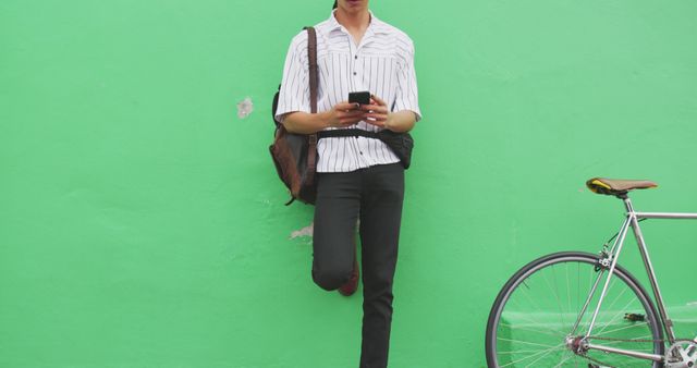 This image shows a young man leaning against a bright green wall while using a smartphone. He has a casual and modern appearance, wearing a striped shirt and holding a bag over his shoulder. A bicycle is resting next to him. This image could be used for themes related to technology, urban lifestyle, youth culture, and communication. It is perfect for illustrating content in blogs, social media, and advertisements targeting young adults.