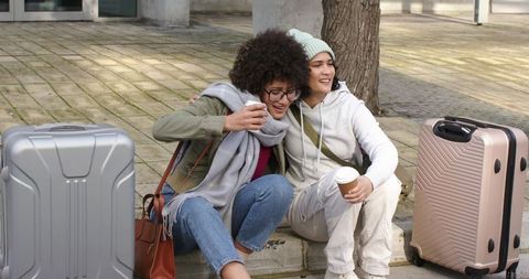 Two young friends sitting on curb holding coffee near suitcases urban travel moment