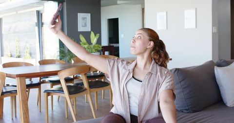Smiling woman taking selfie on gray sofa in modern open-plan living room